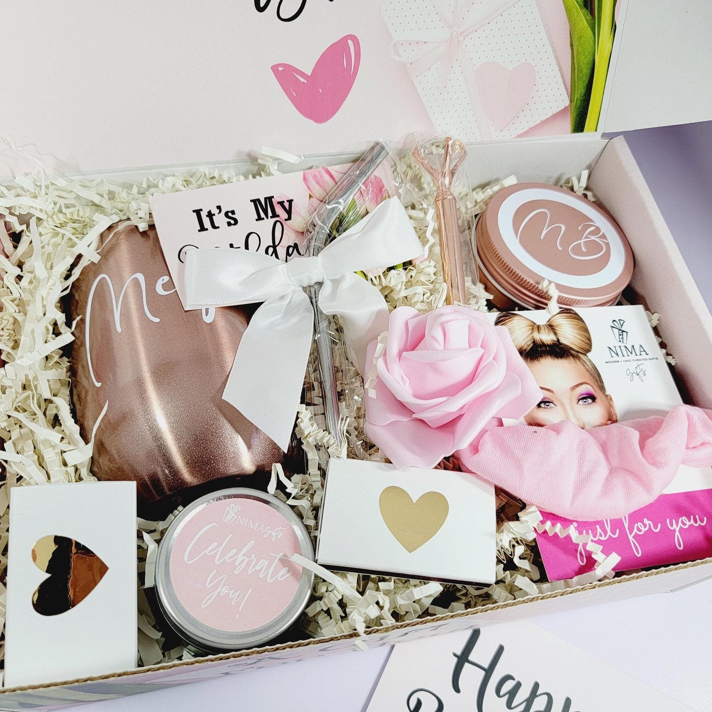 a beautiful and personalized gift box contains a rose gold wine tumbler with a metal straw and a white satin bow, a rose gold candy jar with initials on it, a pink cotton hair scrunchie, a small ethereal lavender candle with rose petals, a box of matches with a gold heart on it, and a rose gold bling pen with a large greeting card that says "Happy Birthday"