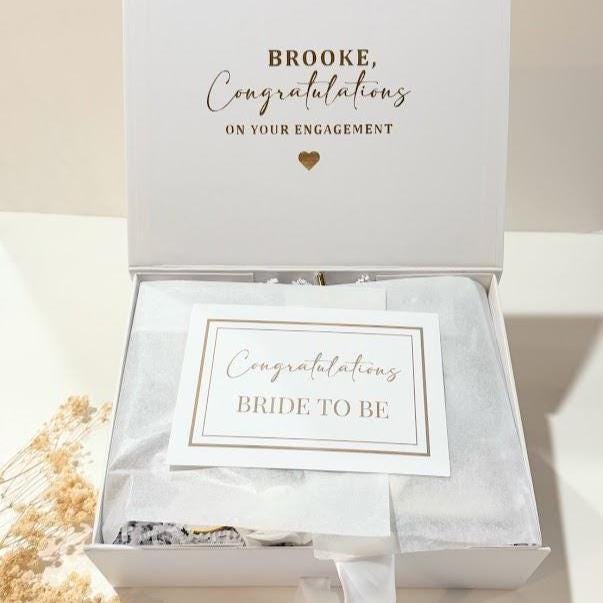 A white magnetic gift box  featuring gold text that reads BROOKE, Congratulations ON YOUR ENGAGEMENT with a small gold heart. Inside, white tissue paper covers the contents, topped with a card reading Congratulations BRIDE TO BE.