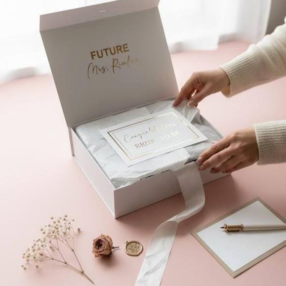 This Personalized Bride to Be Engagement Gift Box is filled with a  personalized coffee mug, personalized bride champagne tumbler with a gold metal straw and personalized satin bow, personalized ring dish, a white satin hair scrunchie and a white foam rose. A greeting card to make your wishes and ask packed in a white magnetic keepsake gift box personalized just for her.