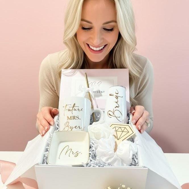 This Personalized Bride to Be Engagement Gift Box is filled with a  personalized coffee mug, personalized bride champagne tumbler with a gold metal straw and personalized satin bow, personalized ring dish, a white satin hair scrunchie and a white foam rose. A greeting card to make your wishes and ask packed in a white magnetic keepsake gift box personalized just for her.
