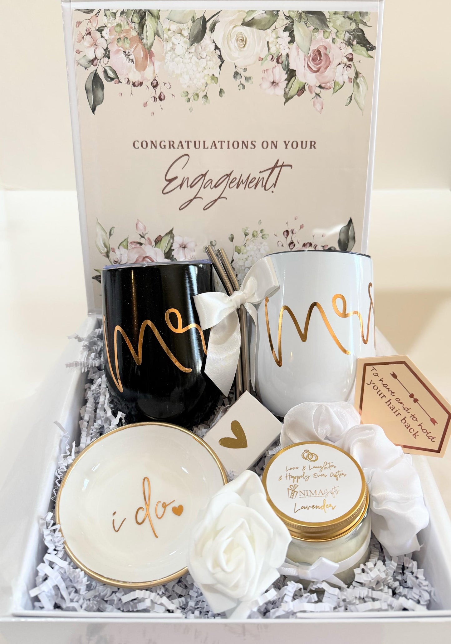 A gift box filled with wedding-themed items, including two black and white mugs with the words "Mr." and "Mrs." written in gold script, a small white candle with the words "Love & Laughter & Happily Ever After" written in gold script, a white rose, and a white dish with the words "I do" written in gold script.