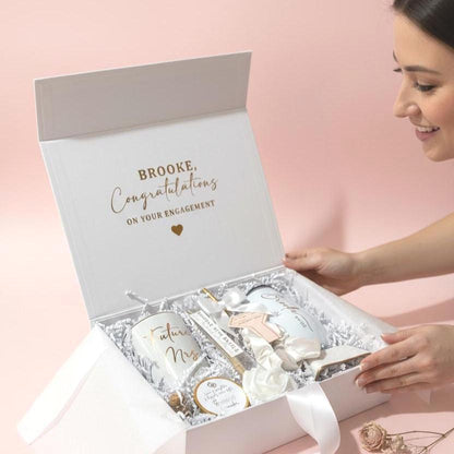 A white gift box filled with wedding-themed items. Includes a mug with "Future Mrs." in gold script, a tumbler with "Elina Wifey", a ring dish, a candle, and a decorative rose. The box is filled with white paper filler.