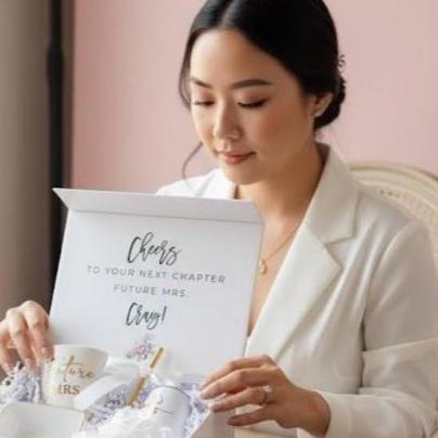 A bride to be is sitting and looking at an open box of the bride engagement gift box that contains a personalized coffee cup with Future Mrs and her last name on it, a white "Bride" 6oz tumbler with a gold straw and white bow, a white ring dish with gold trim and the word "Mrs" on it, a white hair satin hair scrunchie and a white rose. All packaged in a "keepsake" personalized with her first name magnetic gift box. Great gift for an engagement party.