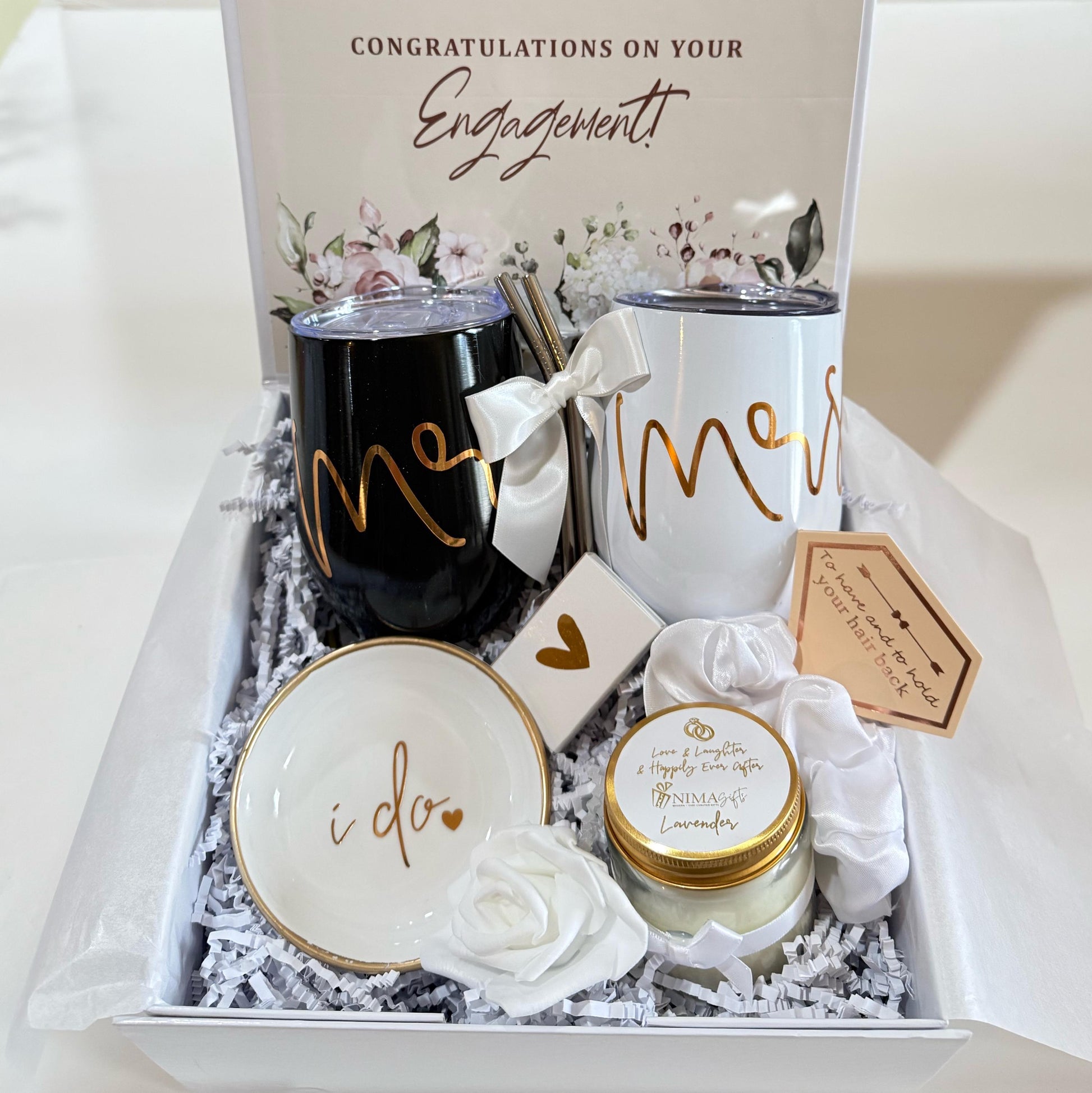 A gift box filled with wedding-themed items, including two black and white mugs with the words "Mr." and "Mrs." written in gold script, a small white candle with the words "Love & Laughter & Happily Ever After" written in gold script, a white rose, and a white dish with the words "I do" written in gold script.