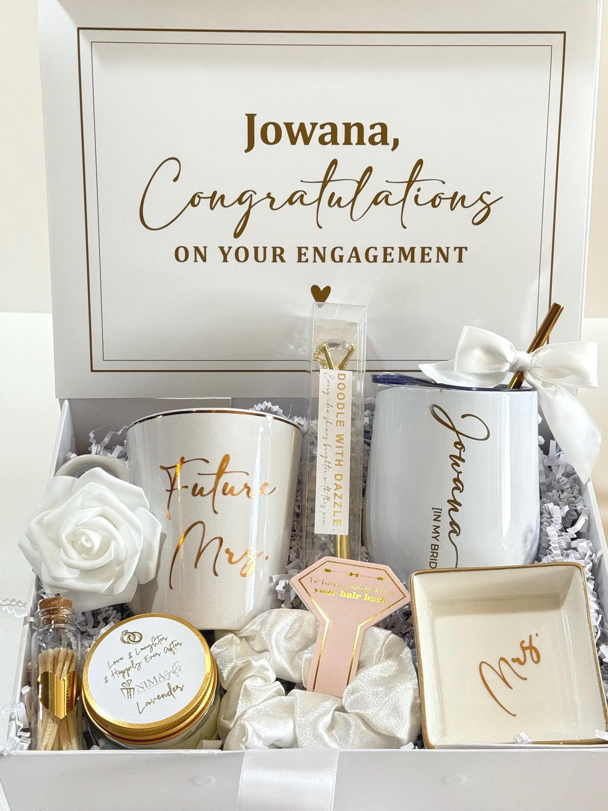 A white gift box containing a personalized mug with "Future Mrs. Last Name” in gold, a white engraved tumbler with "Jowana" and "IN MY BRIDE ERA" in gold, a white rose, a candle, a white bling pen, a white scrunchie, a personalized ring dish with "FUTURE MRS. LAST NAME.", and a small bottle of matches.