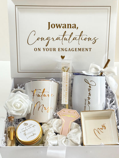 A white gift box containing a personalized mug with "Future Mrs. Last Name” in gold, a white engraved tumbler with "Jowana" and "IN MY BRIDE ERA" in gold, a white rose, a candle, a white bling pen, a white scrunchie, a personalized ring dish with "FUTURE MRS. LAST NAME.", and a small bottle of matches.