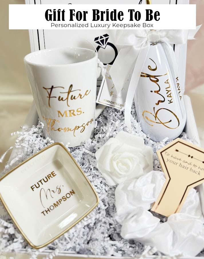This Personalized Bride to Be Engagement Gift Box is filled with a  personalized coffee mug, personalized bride champagne tumbler with a gold metal straw and personalized satin bow, personalized ring dish, a white satin hair scrunchie and a white foam rose. A greeting card to make your wishes and ask packed in a white magnetic keepsake gift box personalized just for her.