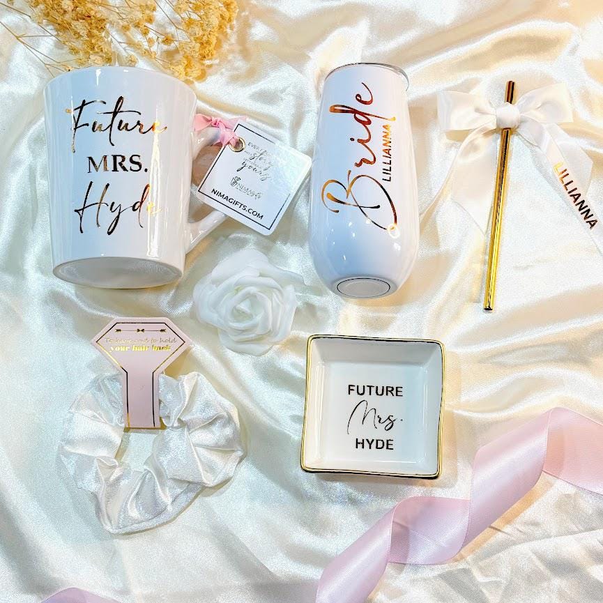 This Personalized Bride to Be Engagement Gift Box is filled with a  personalized coffee mug, personalized bride champagne tumbler with a gold metal straw and personalized satin bow, personalized ring dish, a white satin hair scrunchie and a white foam rose. A greeting card to make your wishes and ask packed in a white magnetic keepsake gift box personalized just for her.