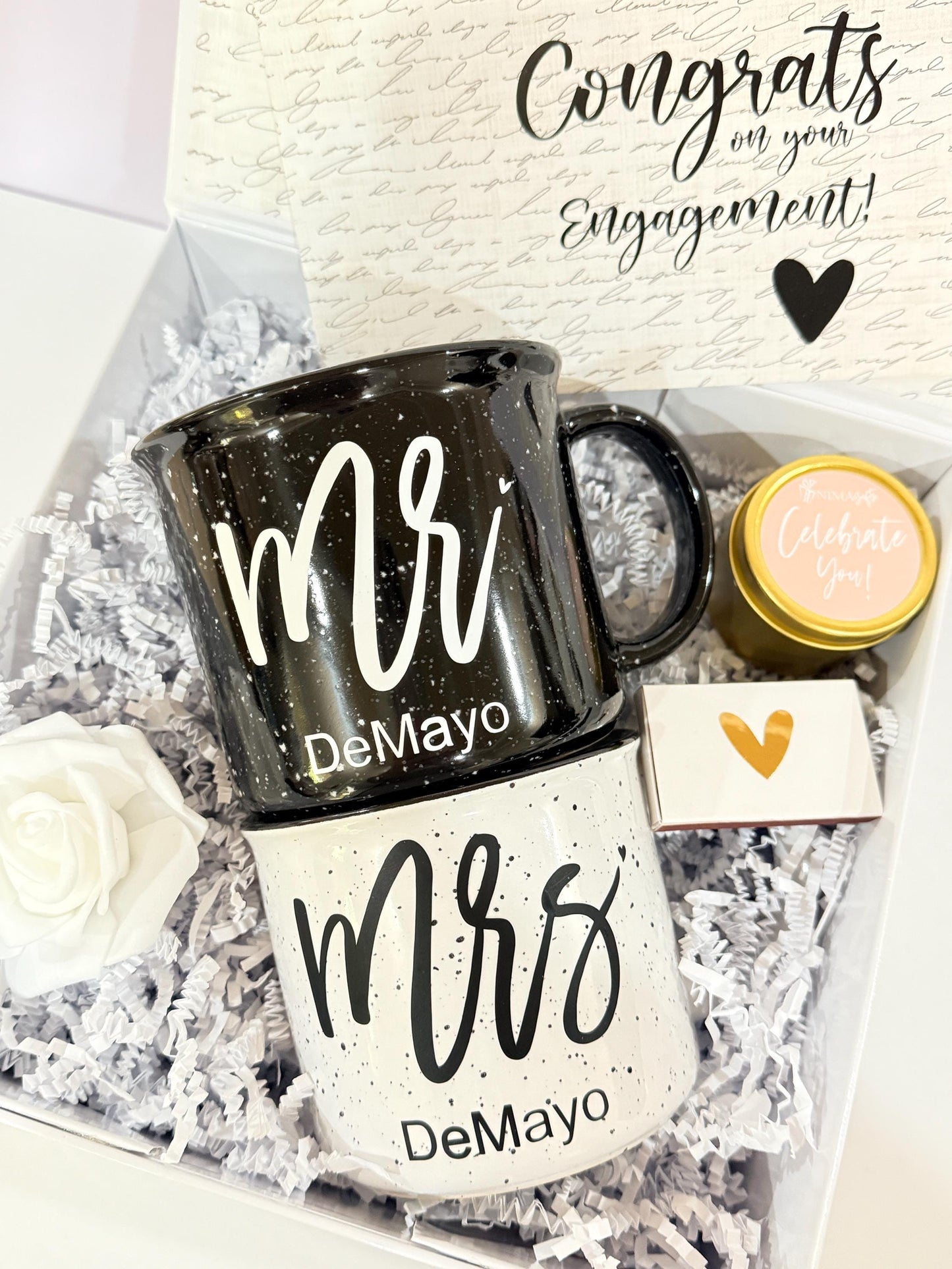This engagement gift box for couple is personalized with the couple last name, the mr and mrs ceramic campfire mugs in black and white with a hand poured lavender candle and a box of matches are packed in a white magnetic box with personalization on the front and inside the gift box as well as the mugs. A matching greeting card is also visible in the image and is included in the gift box.
