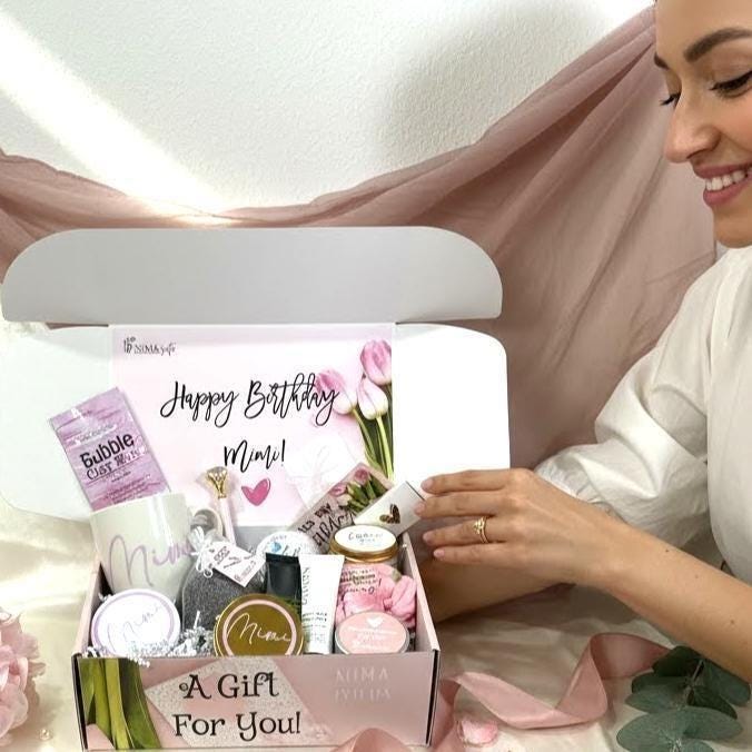 Spa birthday gift box for women that includes a personalized coffee mug, initialed leatherette compact mirror and an initialed candy jar, a bath hand made bath bomb, body lotion, body butter, a hand poured lavender candle and matches, a cotton hair scrunchie, a sachet with lavender leaves and a travel size bar of black soap. Packaged in a beautiful personalized tulip gift box.