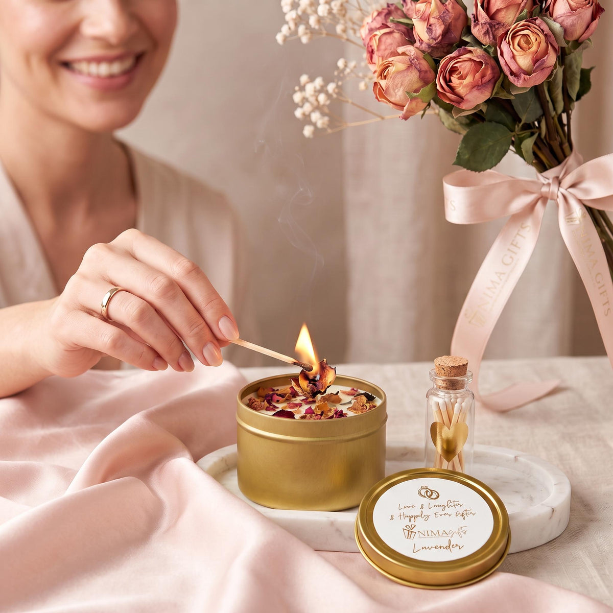 bride to be is lighting a hand poured lavender candle with rose petals inside a gold tin. Top of candle reads “ Love and laughter, and happily ever after” in gold letters. A small glass bottle filled with matches with white tops, and a gold heart on top of it is also included in the image.