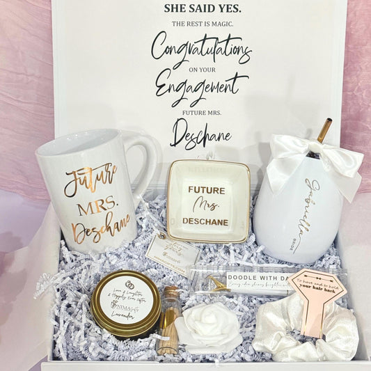 A beautiful bride to be magnetic white box is filled with a 15 ounce mug that says future misses and the girls last name it has a ring dish made that a porcelain with gold trim and gold letters inside to say future Mrs. and the girls‘s last name, White Tumblr engraved with the girls first name a gold medal a straw and a satin bow also comes with a 4 ounce hand poured candle matches in a white in a glass box. A white rose, a white pen with bling and gold trim, and a white satin scrunchie .