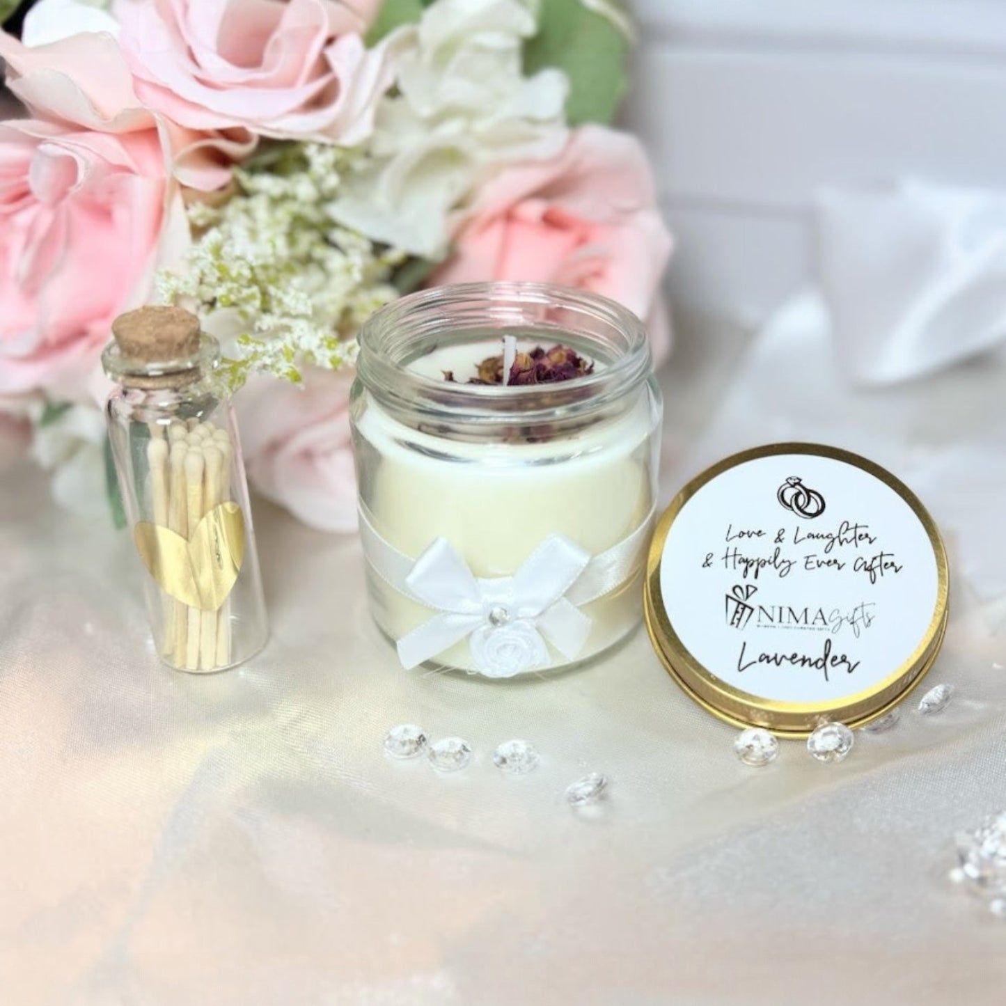 NIMA Gifts lavender scented soy candle with gold lid, white ribbon, and rose petals, next to a glass vial of matches with a gold heart.