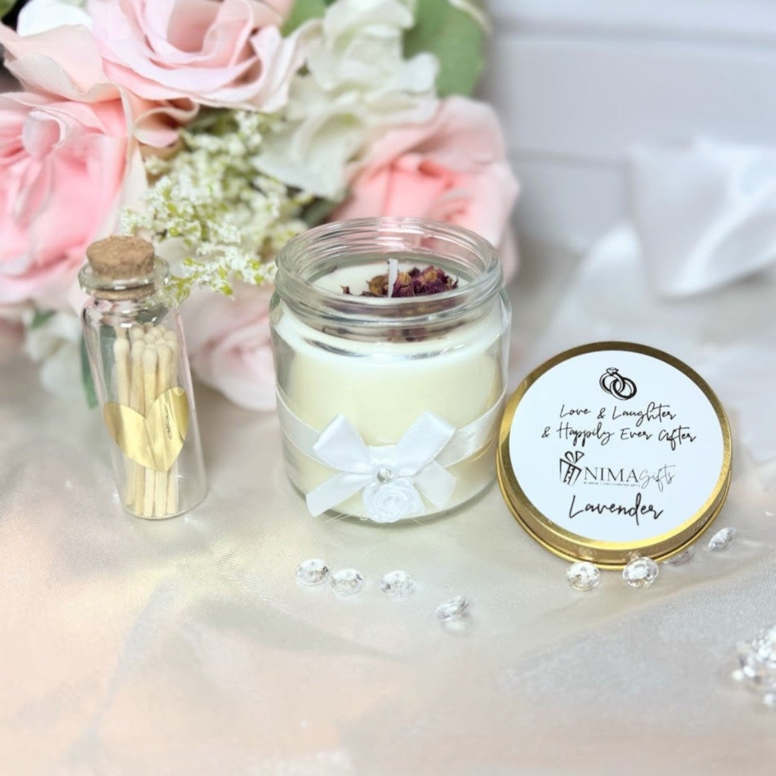 NIMA Gifts lavender scented soy candle with gold lid, white ribbon, and rose petals, next to a glass vial of matches with a gold heart.