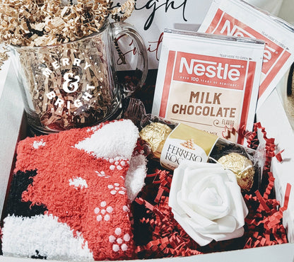 A personalized Christmas gift box with a white mug, red socks, white and red Christmas candies, a note card, and a red foam rose, all presented on shredded paper.