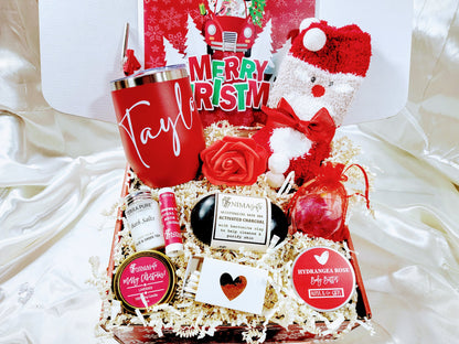 A Christmas gift box for women, containing a variety of holiday-themed items such as a red wine tumbler, bath bomb, lip balm, socks, a notecard, and a body butter, all arranged in a decorative manner with holiday decorations.